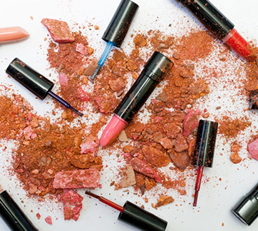 Scattered and crushed makeup products, including lipsticks and powder in shades of pink and brown, on a white surface.