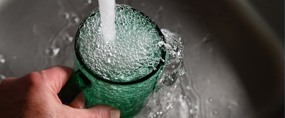 A hand holds a green glass mug as a stream of water fills it, creating bubbles and overflowing into a sink.